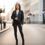 Mladá žena v čiernej koženej bunde stojaca na ulici počas zlatej hodiny, mestské pozadie.