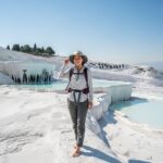 Mladá žena v svetlom oblečení stojí bosá na travertínových terasách Pamukkale, Turecko, pri slnečnom dni.